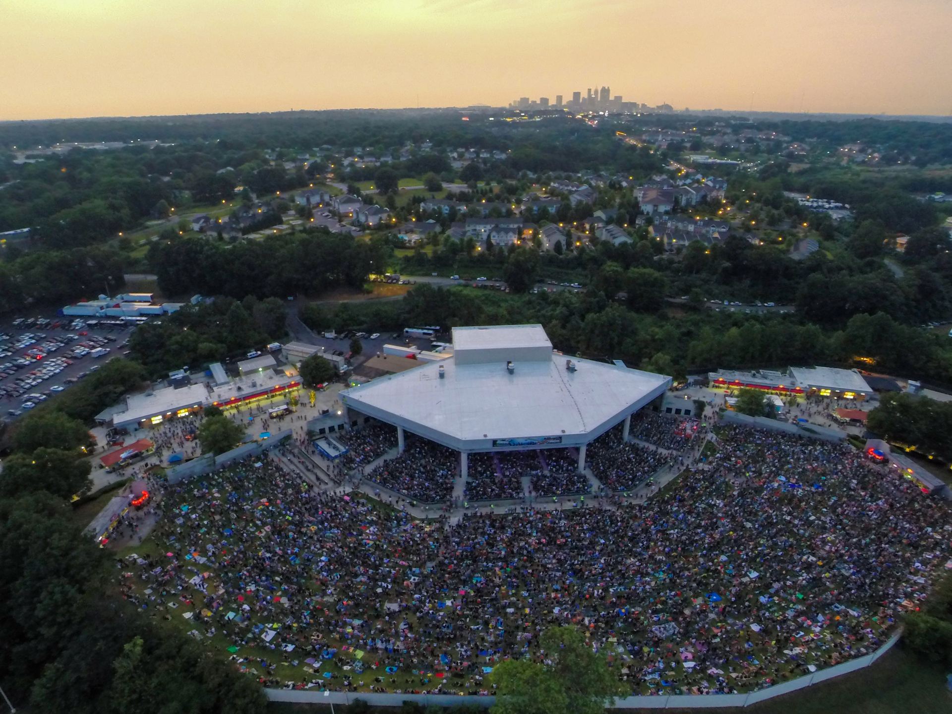 Aerial view at dusk of a packed concert at Lakewood Amphitheatre, with thousands of fans on the lawn and the Atlanta skyline on the horizon